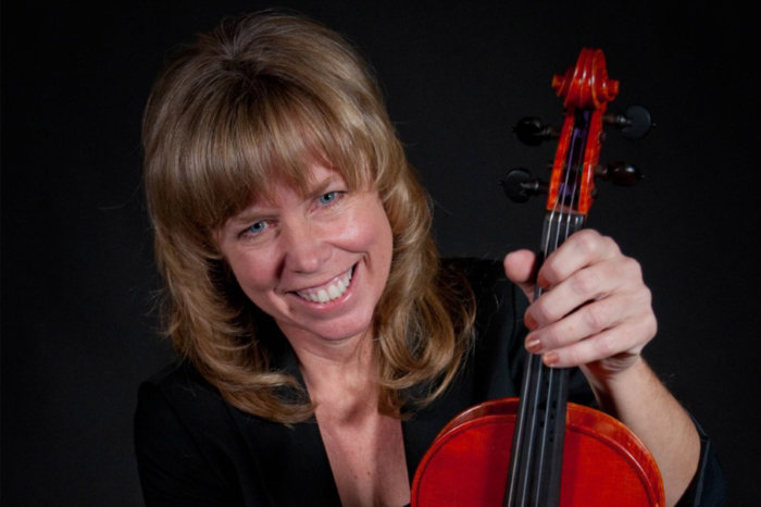 Headshot of Lorie Hippen. She is wearing a black outfit against a black background. She is smiling and holding a violin. She has long hair with bangs.