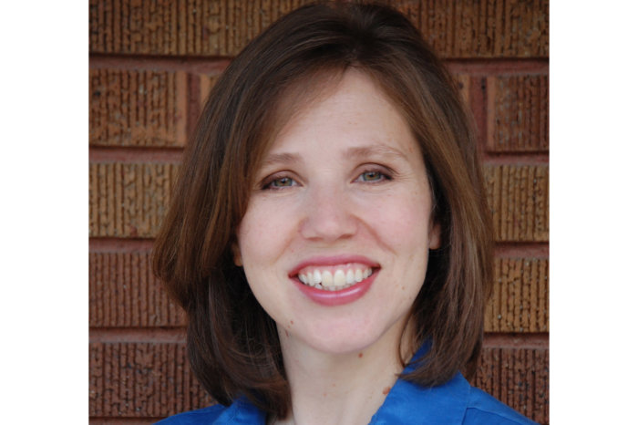 Headshot of Maria Jones. She has short brown hair and is smiling. She wears a blue shirt and is standing in front of a brick wall.