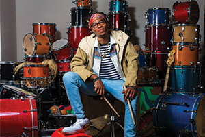 LA sitting on a stool with a bunch of drums behind him