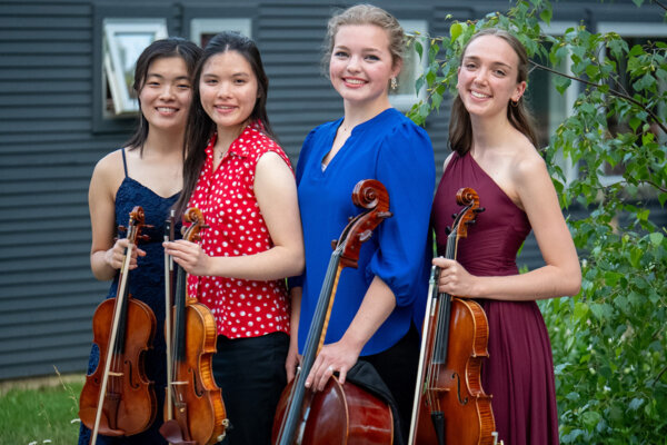 Four string players posing with their instruments in a rustic setting