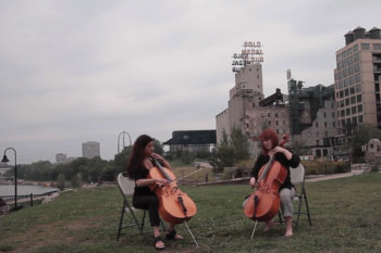 Two people playing cello outside with Minneapolis skyline and Mississippi river behind them