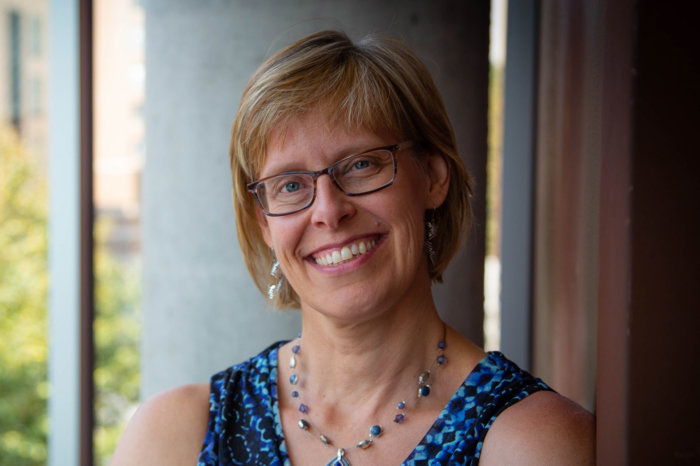 Headshot of Sue Ruby. She has short hair and wears glasses. She is wearing a blue shirt and blue necklace.