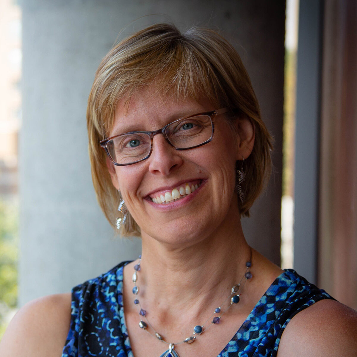 Headshot of Sue Ruby. She has short hair and wears glasses. She is wearing a blue shirt and blue necklace.