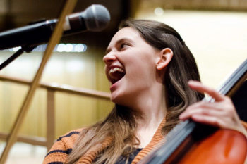 Action shot of Carlynn Savot from the shoulders up. She is playing the cello and smiling in front of a microphone.