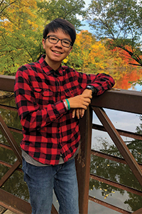 Young student wearing a red and black flannel shirt standing outside on a bridge over water.