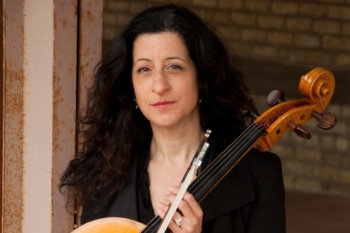 Headshot of Jacqueline Ultan. She is wearing a black shirt and has black curly hair. She is holding a cello in front of a brick wall.