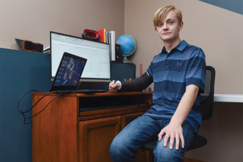 Young student wearing a blue polo shirt and jeans, sitting a desk with a computer sitting on it.