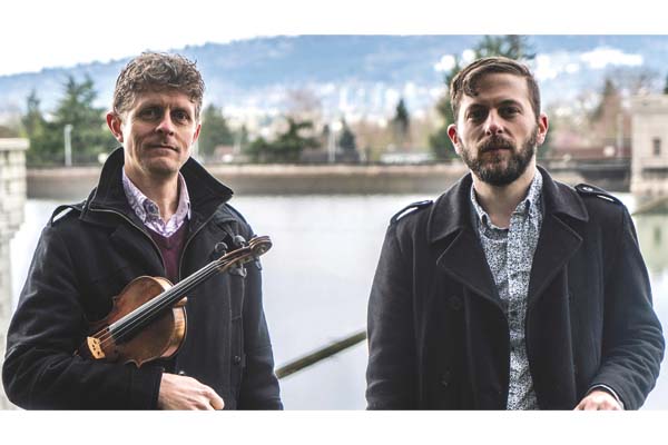 Two people standing next to each other outside. One is holding a violin and the other is holding a guitar