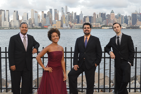 4 adults standing in front of a body of water and the New York City skyline