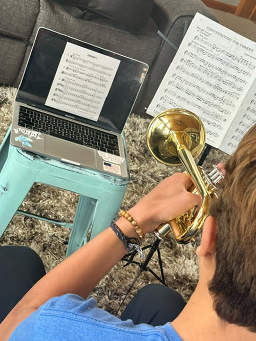 Student playing trumpet in front of a computer screen doing online instruction
