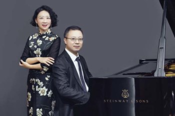 Two people at a piano. One person is sitting and the other is standing behind them.