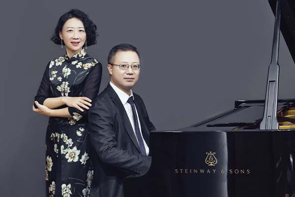 Two people at a piano. One person is sitting and the other is standing behind them.