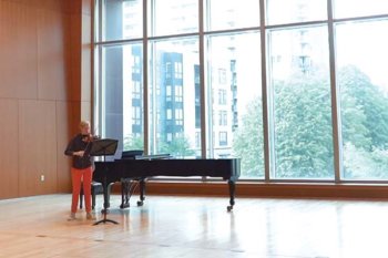 Person playing a violin. They are standing next to a violin by floor to ceiling windows.