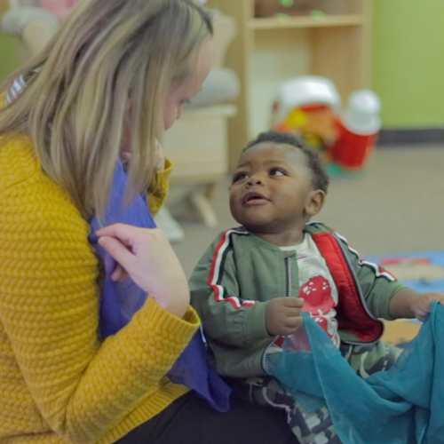 Teacher looking at a baby. Both are holding scarves